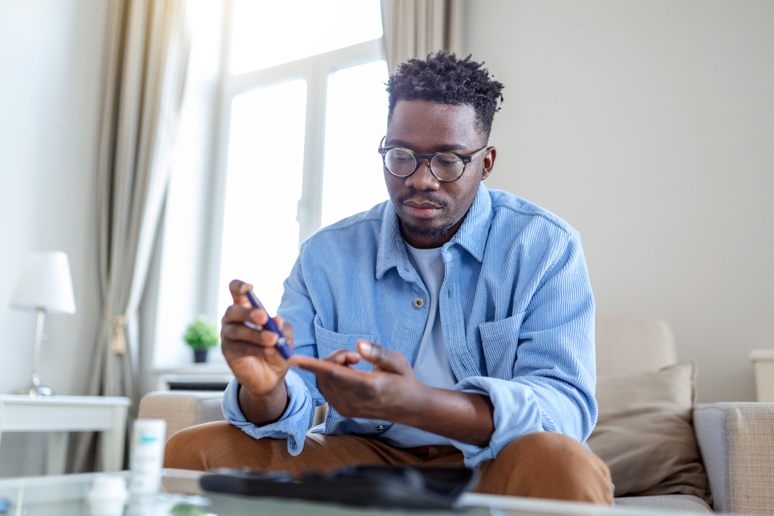 African man is sitting at the sofa and taking blood from his finger due to diabetes. The daily life of a man of African-American ethnicity person with a chronic illness who is using glucose tester.